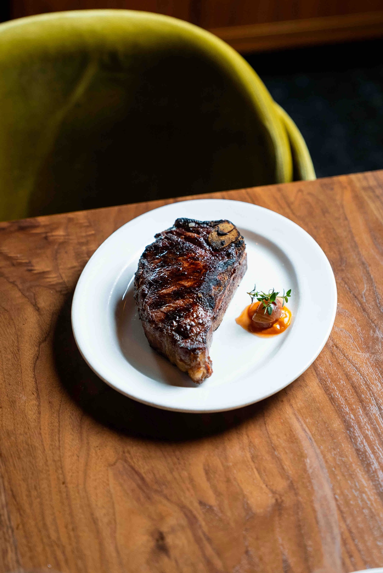 A char-grilled bone-in ribeye on a white plate beside a green velvet chair — the wood-fired cuts at The Arthur J