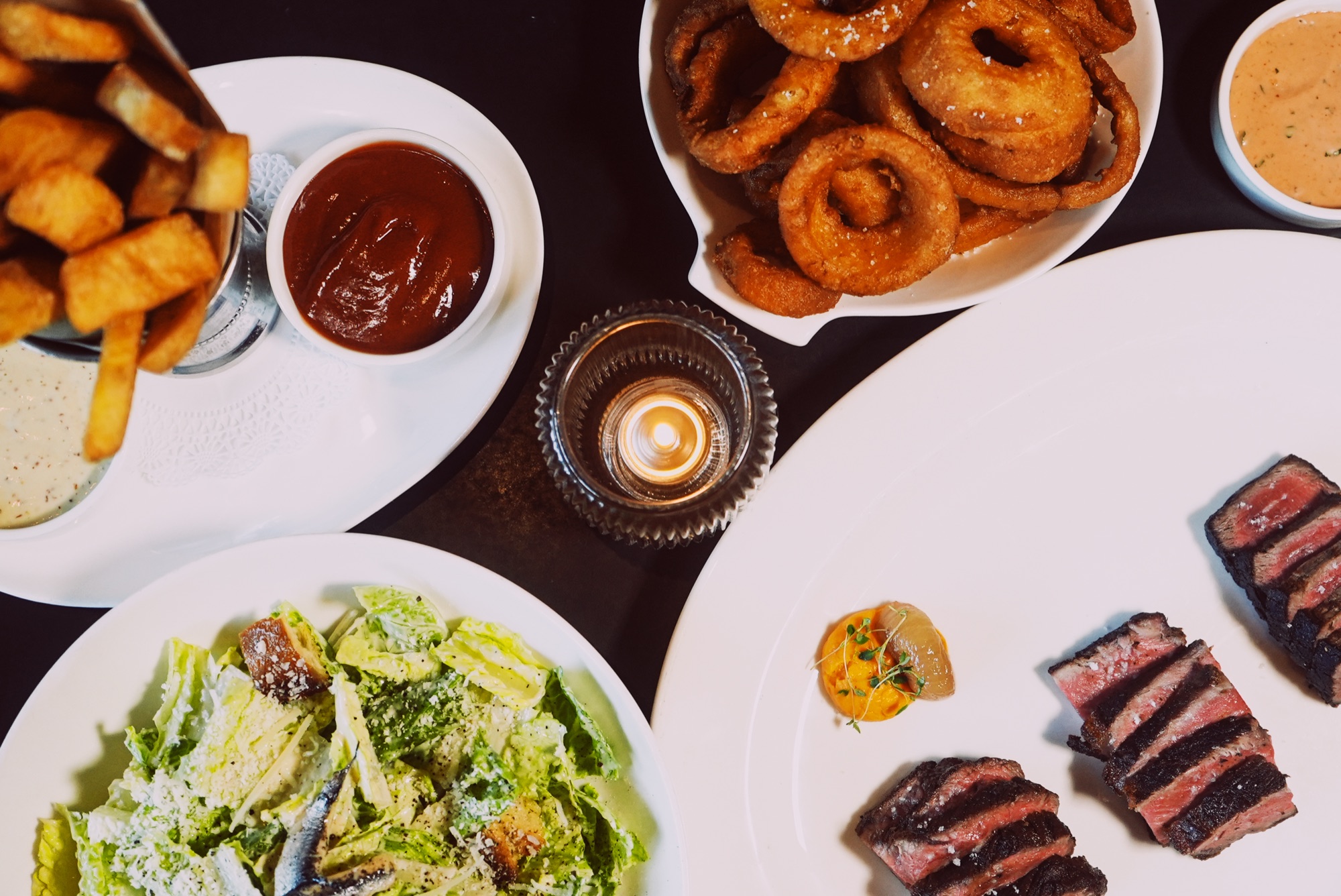 An overhead candlelit spread of sliced steak, hand-cut fries, onion rings and a Caesar salad on a dark booth table at The Arthur J