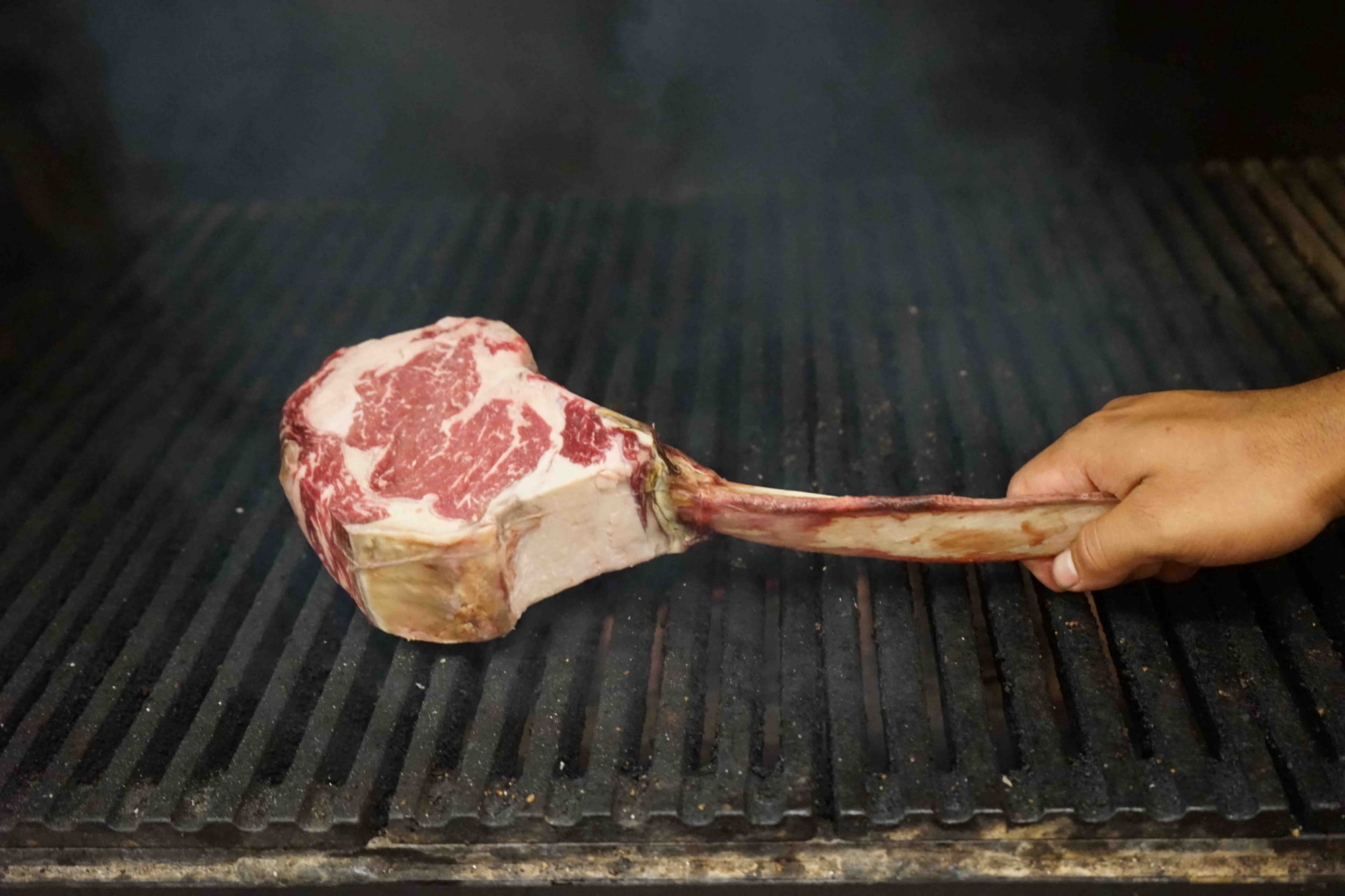 A chef placing a marbled bone-in tomahawk steak onto the live wood-fire J&R grill at The Arthur J